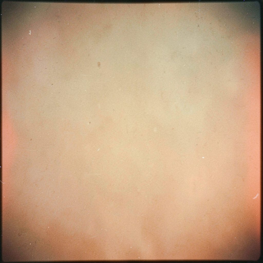 Soft analog film photograph with visible grain, warm cream and muted lavender tones, gentle domestic still life with fabric folds and a window casting pale light.